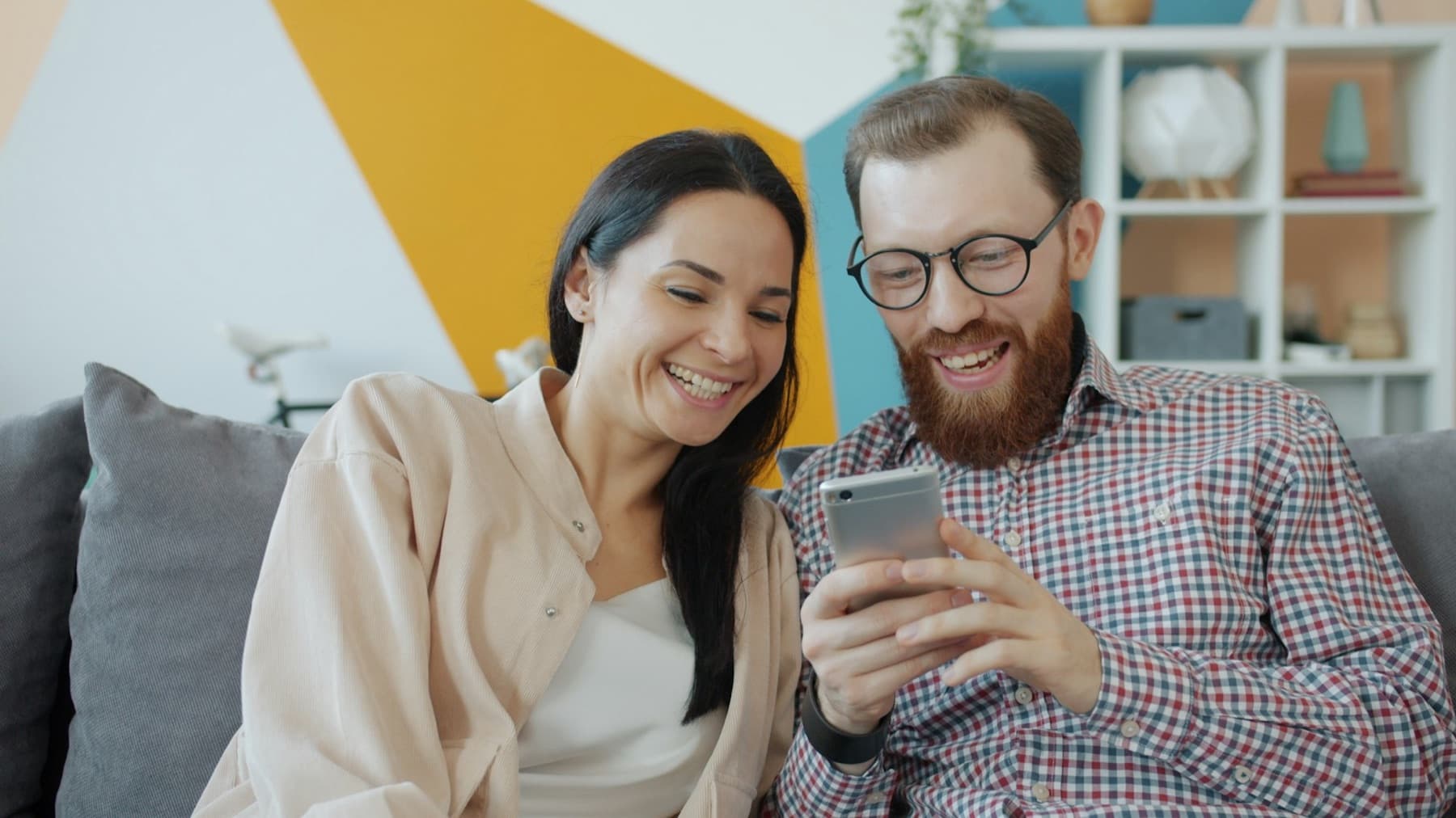 A couple laughing at a phone on a grey couch in front of a colorful wall