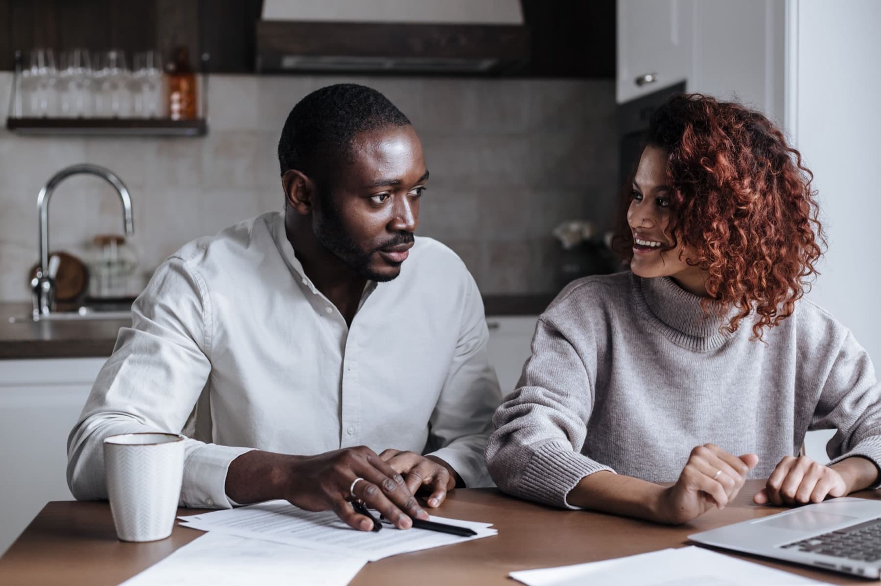 A couple smiling at each other over papers and a laptop at the kitchen table
